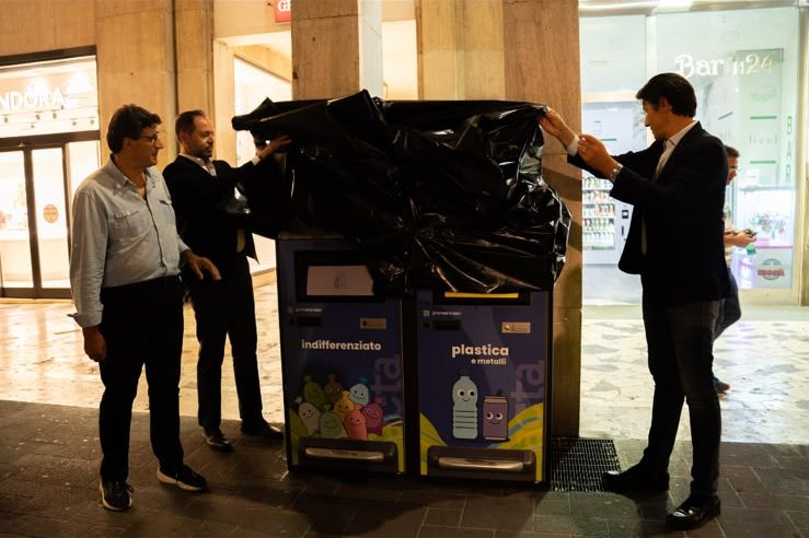 Tre persone scoprono i nuovi cestini smart compattanti installati in strada: due contenitori affiancati, uno per l'indifferenziato e uno per plastica e metalli.